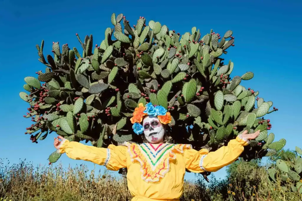 Person in yellow dress with Día de los Muertos makeup posed before nopales cactus